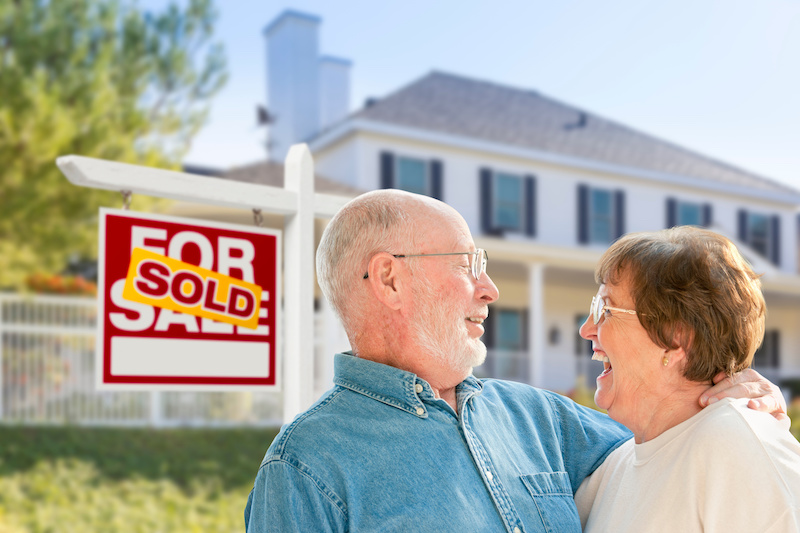 Happy Affectionate Senior Couple Hugging in Front of Sold Real Estate Sign and House.